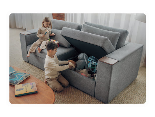 Two children playing beside a charcoal Rezy Smart Sofa with the under-seat hidden storage compartment open and full of toys
